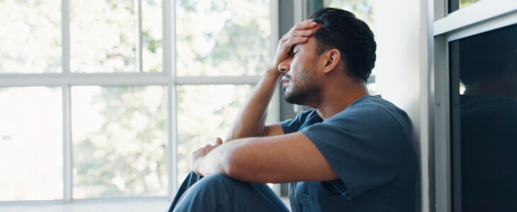 Exhausted man in scrubs sitting by window with hand on forehead