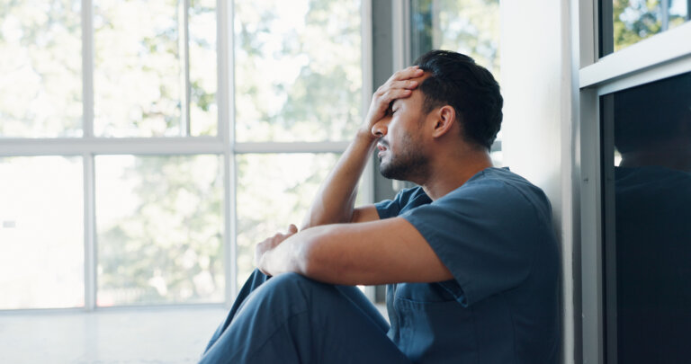 Exhausted man in scrubs sitting by window with hand on forehead