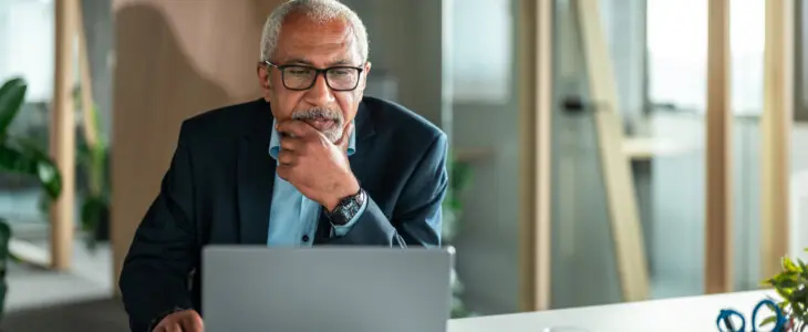 Older man in suit working thoughtfully on laptop at office desk